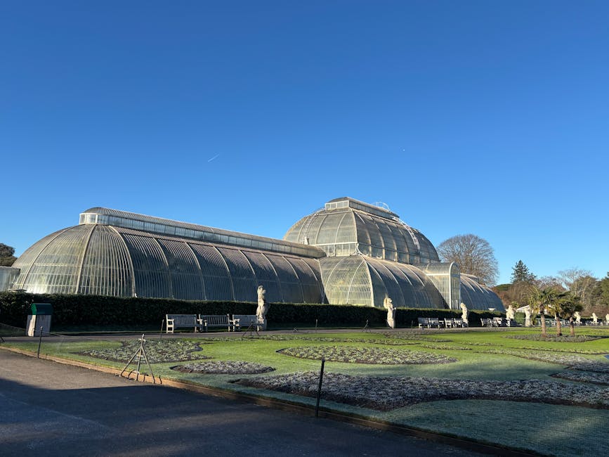 View of a large Victorian glasshouse or conservatory, built with extensive glass panels and decorative white framework, partially obscured by surrounding green trees. The structure appears to be part