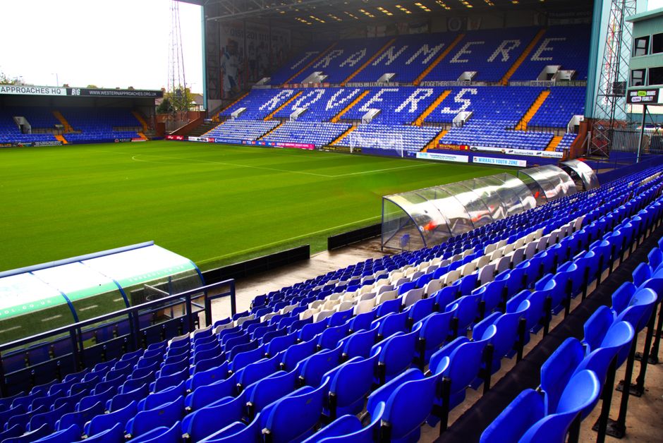 Inside a football stadium with a well-maintained grass playing field and rows of blue plastic seats arranged in ascending tiers, adjacent to a covered team bench area with transparent plastic panels s